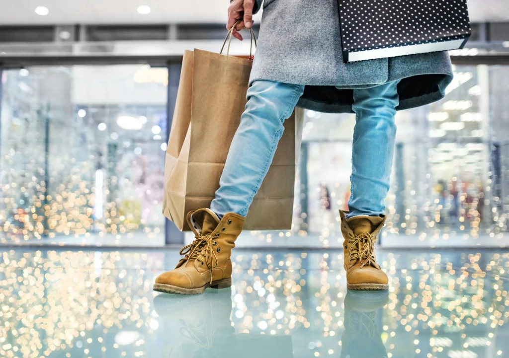 Chica sujetando una bolsa de compras, solo se ve de su cintura hasta bajo.
LLeva una blusa comprida gris, pantalones jeans y botas marrón claro